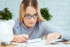 A person with glasses sits at a table making notes while counting coins.