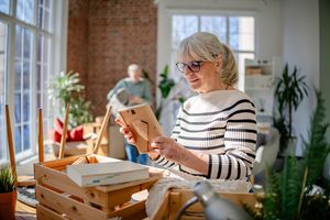 Older woman examining a frame indoors, with individuals and plants in the background, a bright and airy room
