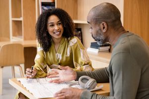 Two people at a table discussing a blueprint, one appears engaged and smiling while holding a pen