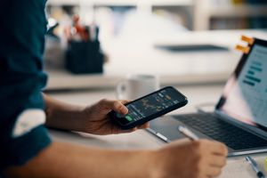 A man looks at financial information on a smart phone while sitting in front of a laptop.