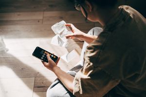 A person sits on the floor, using a calculator on their smartphone while reviewing receipts. 