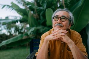 An older man wearing glasses, sitting outdoors with a pensive expression and lush green plants in the background