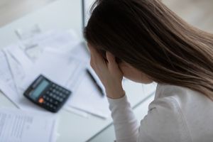 A person sitting at a desk with documents holding their head in their hands near a calculator