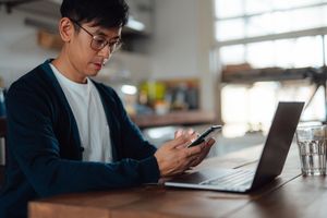 A young investor checks on investments using a phone and a laptop at a kitchen table.
