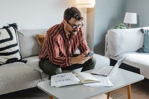 A man seated on a sofa using a smartphone with a laptop and papers on the table in front of him