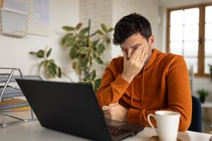 Man sitting at a desk with a laptop covering his face with his hand appearing stressed