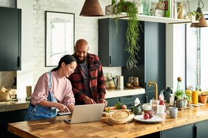 Two people using a laptop in a modern kitchen while preparing food smiling and enjoying their time together