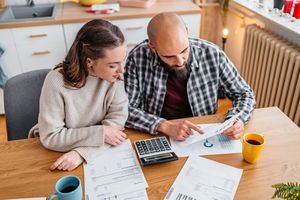 Two individuals seated at a table reviewing financial documents with a calculator cups and papers in a home setting