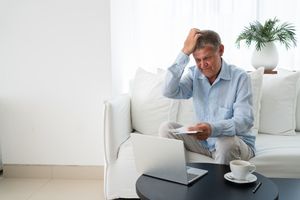 Man sitting on a couch holding a paper and scratching his head, a laptop and coffee on the table in front of him