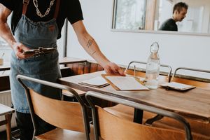 A man wearing an apron works in a small, artisan cafe. He picks up menus and places them on a table.