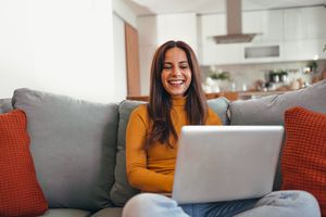 A cheerful woman sits comfortably on her couch using a laptop, enjoying a relaxing day at home in her cozy living room.