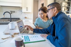 Two people working together at a laptop in a kitchen with papers on the counter