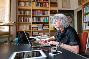 An older woman working on a laptop at a desk in a home office setting with bookshelves in the background