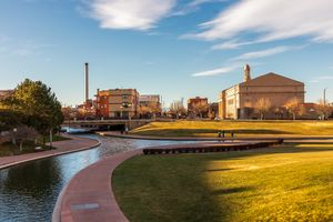 A canal and grasslined walkway in an urban park setting with buildings and a smokestack in the background