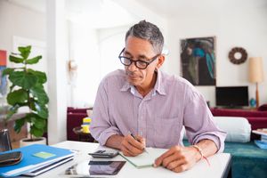 An investor seated at a table in his living room uses a pen, pad, calculator, and mobile device to determine how much he has available to allocate to his retirement portfolio.