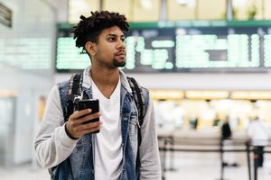 A person standing in an airport holding a smartphone arriving or departing terminal background with flight information displays