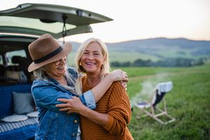 Two cheerful older women hugging outside a car with a scenic background of fields and hills