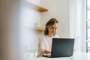 A female investor uses a laptop computer to manage her investments while sitting at a kitchen island. 