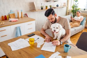 A couple chilling in their living room with the dog sitting with one filling paperwork while the other looking at tablet on the sofa.