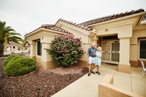 An older couple standing in front of a house with a tiled roof and landscaped surroundings holding hands and smiling at each other