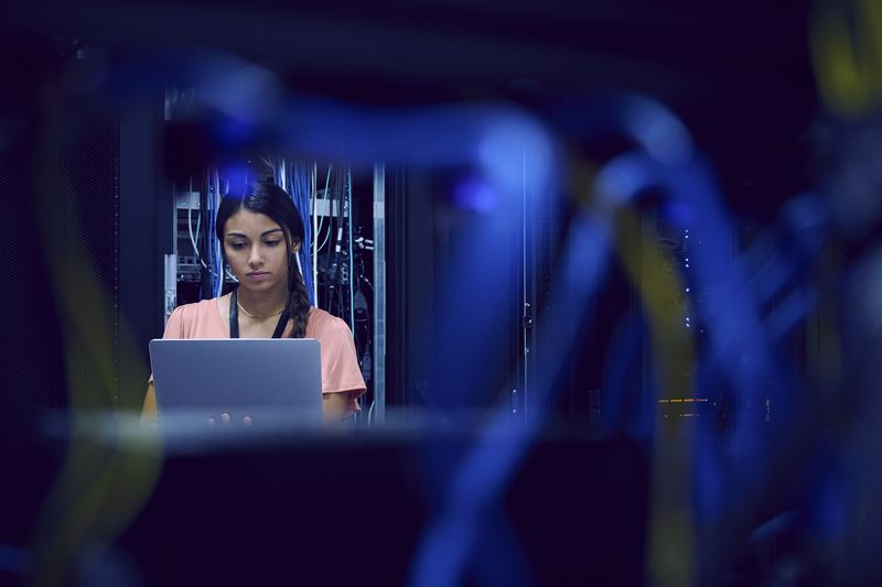 A person working on a laptop in a server room with network cables in the foreground