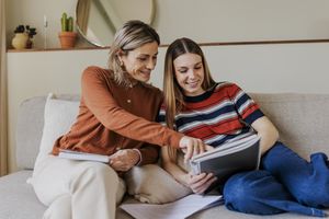Two women sitting on a couch one pointing at a notebook while they both look at it