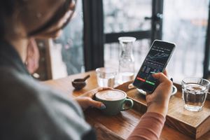 Person holding a coffee cup and viewing a stock trading app on a smartphone seated at a wooden table
