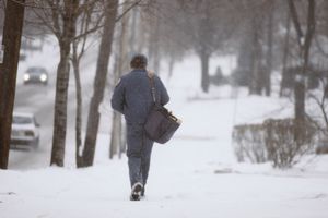 A mail carrier walking on a snowy sidewalk while carrying a shoulder bag
