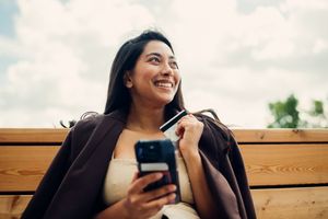 A person holding a smartphone and smiling while sitting outdoors on a wooden bench