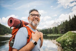 A man outdoors wearing a backpack and smiling in front of a forested lake scene