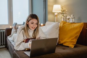 Young woman on her sofa looking happily at her laptop with holiday lights in the background