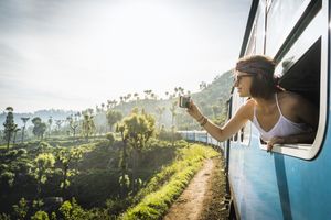 Person leaning out of a train window holding a phone and taking a photo of the scenery with trees and tracks