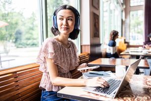 A woman sitting at a laptop with headphones on holding a phone in a caf
