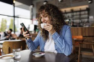 A person holding a phone and drinking from a cup in a cafe