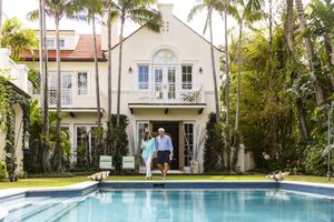 Two people walking by a swimming pool in front of a large house with palm trees