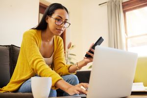 A woman holding a phone while using a laptop at home
