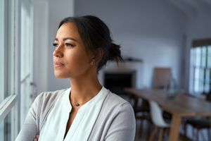 A thoughtful person standing by a window arms crossed looking outside