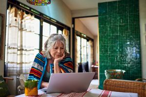 A person wrapped in a blanket sits at a table focusing on a laptop in a cozy room with a green tiled wall
