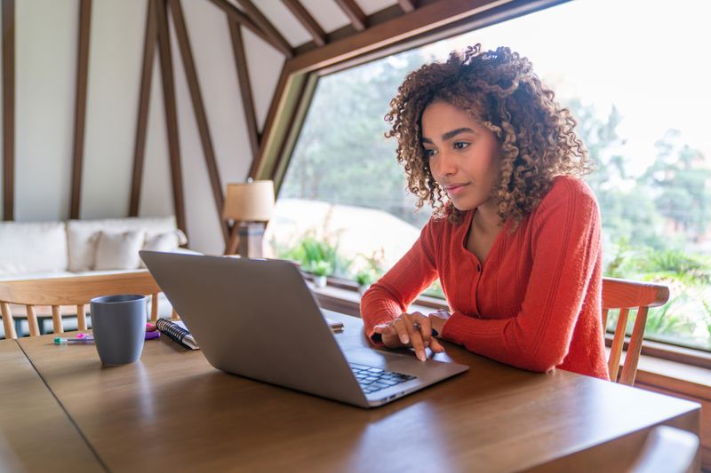 A person working on a laptop at a wooden table in a cozy welllit room with a triangular window in the background