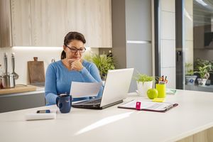 A person sitting at a kitchen counter working on a laptop holding a piece of paper with a thoughtful expression