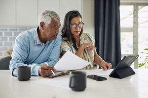 An elderly couple is sitting at their kitchen counter reviewing their finances. They have a tablet and paperwork in front of them.