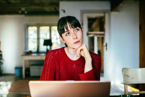 Woman around age 30 sitting at home and wondering while sitting in front of her laptop