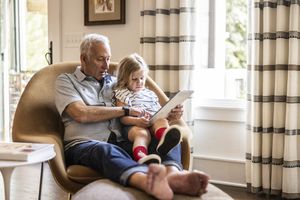 An older man sitting with a young girl on his lap reading a tablet together in a comfortable living room setting.