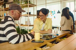 Two people sitting at a table having a discussion with additional individuals conversing in the background of a workspace