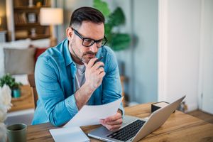 Man in his mid-30s sitting at home and looking seriously at documents and a laptop