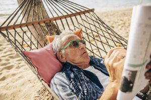 An older person reading a newspaper while lying in a hammock on a beach
