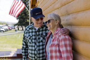 Two older adults dressed casually the man has his arm around the woman they are outdoors next to a log building with an American flag visible