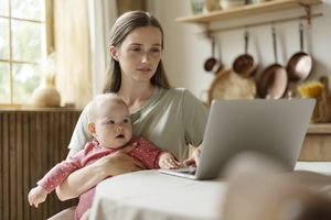 Young mom at home, looking seriously at her laptop with a baby on her lap