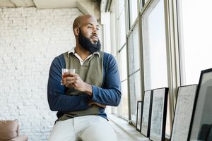 A man holding a drink looking out of a window while sitting in an apartment