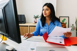 A person sitting at a desk working on a computer holding papers and looking at the screen